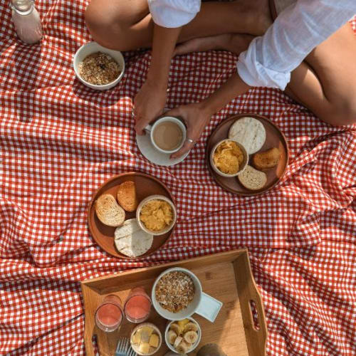 Eine Person sitzt auf einer rot-weißen karierten Decke mit Frühstücksartikeln, darunter Toast, Müsli, Bananen und Getränke, während eines Picknicks oder Campings.