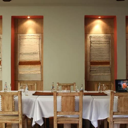 A dining table set with a white tablecloth, wooden chairs, and water bottles, against a wall with four framed textile art pieces illuminated by spotlights.