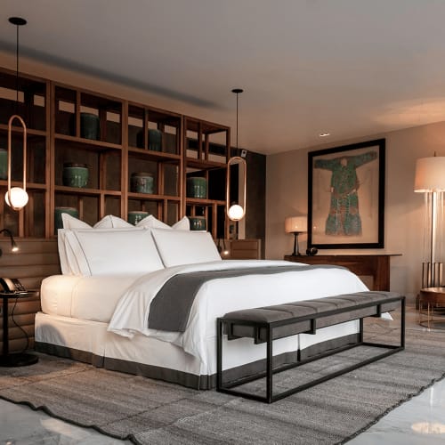 Modernes Hotelzimmer mit einem großen Doppelbett mit weißen Bettwäsche, einer dunklen Bank am Fußende des Bettes und Holzregalen dahinter, die grüne Keramikvasen zeigen, samt ambienten Hängeleuchten und stilvollen Möbeln im Hintergrund.