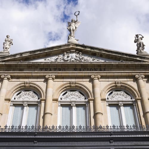 Die verzierte Fassade eines historischen Gebäudes mit klassischer Säulenordnung und Skulpturen auf dem Dach vor einem teils bewölkten Himmel.