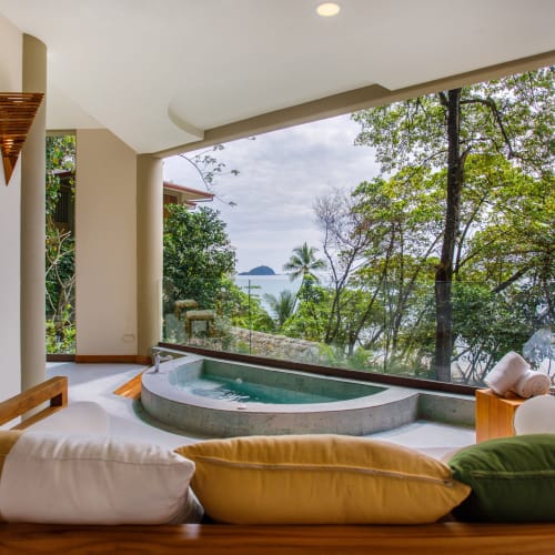 Blick von einem Suitenbalkon mit rundem Whirlpool und Holzmöbeln mit bunten Kissen auf üppige Bäume und eine entfernte Meeresansicht bei bewölktem Himmel.