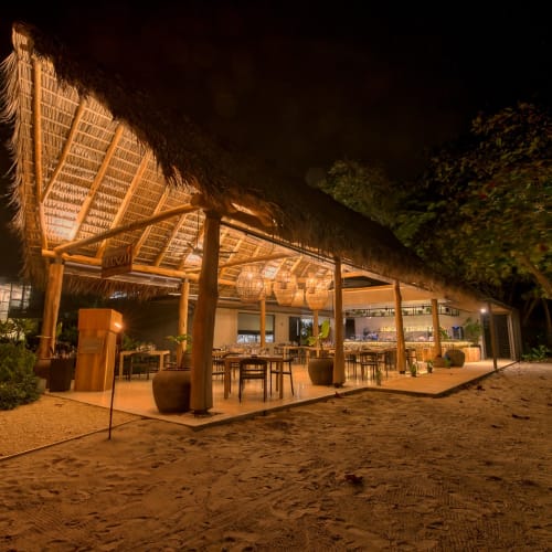 Open-Air-Restaurant mit Strohdach, beleuchtet in der Nacht, an einem Sandstrand mit Bäumen.