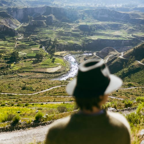 Eine Person mit Hut blickt auf terrassierte Felder und einen Fluss in einer bergigen Landschaft.