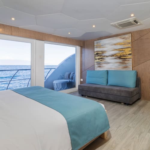 Modern hotel suite with a large bed, teal accents, sofa, and large windows offering a view of the ocean and balcony outside.