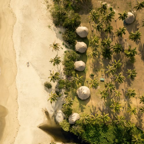Drohnenaufnahme eines Sandstrandes mit Meereswellen links und einer Ansammlung von Palmen sowie mehreren Strohhütten rechts, umgeben von Vegetation.