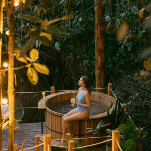 A woman in a blue swimsuit relaxes in a wooden hot tub with a glass of drink, surrounded by dense jungle foliage at sunset.