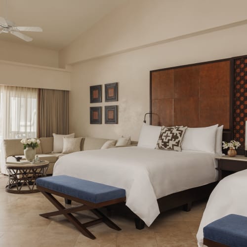 Modernes Hotelzimmer mit zwei Doppelbetten, die mit weißen Bettlaken und dekorativen Kissen ausgestattet sind, einem Sitzbereich mit einem Sofa und einem runden Tisch sowie einem großen Fenster mit Blick auf einen Balkon mit Meerblick.
