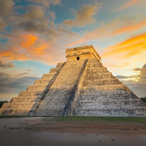 Die Pyramide von Chichen Itza im Licht eines dramatischen Sonnenuntergangshimmels mit verstreuten Wolken, umgeben von Grünflächen.