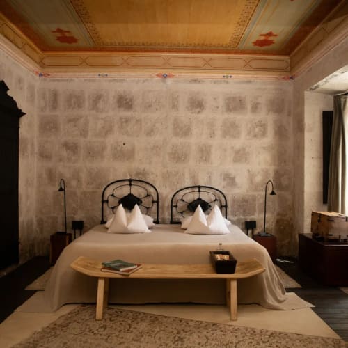 Rustikales Schlafzimmer mit Doppelbett, dekorativen Kissen, Holzbank am Fußende des Bettes und einer bemalten Decke in warmen Farbtönen. Der Raum hat strukturierte Wände, ein Fenster mit Vorhängen und dunklen Holzboden.