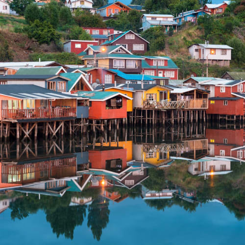 Bunte Häuser auf Stelzen am Seeufer auf der Insel Chiloe, Chile, mit Reflexionen im ruhigen Wasser.