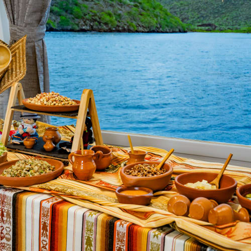 Ein Buffet mit traditionellen Gerichten in Tontöpfen und Holzutensilien auf einem gemusterten Tischtuch in einem Boot mit Fensterblick auf blaues Wasser und grüne Hügel.