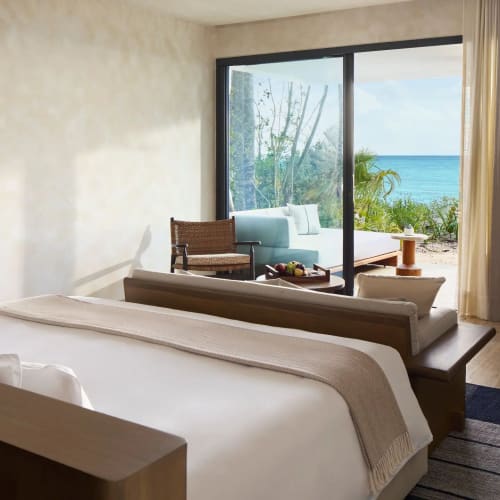 Modernes Schlafzimmer mit großem Bett, hellem neutralem Dekor und Sitzbereich vor bodentiefen Fenstern mit Blick auf den Ozean und eine Terrasse im Freien mit Grünpflanzen.