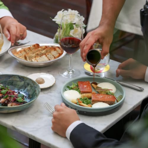Eine Person im Anzug sitzt an einem Marmortisch in einem Restaurant, während ein Kellner Soße über ein angerichtetes Gourmetgericht gießt; eine weitere Person greift nach Besteck, und auf dem Tisch stehen ein Glas Rotwein und ein Brotkorb.