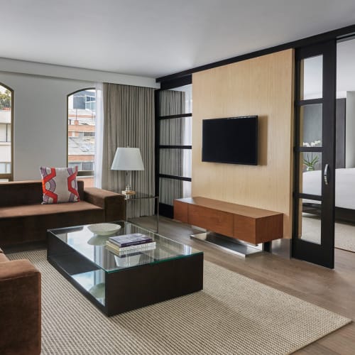 Moderner Wohnbereich mit braunem L-förmigen Sofa mit bunten Kissen, einem Glastisch, einem an der Wand montierten Fernseher auf einer Holzvertäfelung und Blick in ein Schlafzimmer durch Schiebetüren aus Glas in einer Hotelsuite.