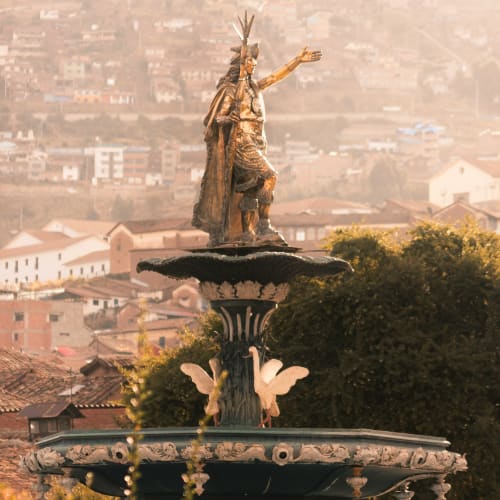 Eine goldene Statue eines sitzenden indigenen Führers mit Federkreuz und ausgestrecktem Arm, auf einem Brunnen mit Stadthäusern und Bäumen im Hintergrund.