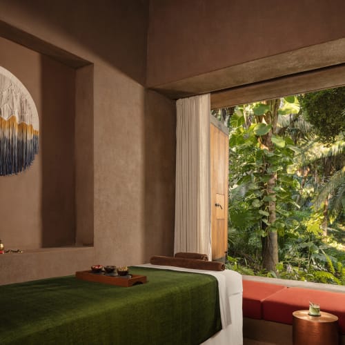 Gemütlicher Spa-Massageraum mit einem Bett, das mit einer grünen Decke bedeckt ist, einem hölzernen Tablett mit Massageutensilien und einem großen offenen Fenster, das eine üppige tropische Vegetation draußen zeigt.