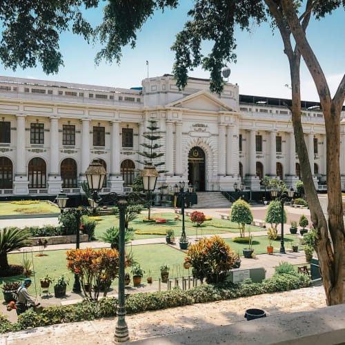 A grand white colonial-style building surrounded by a well-maintained garden and tall trees in Lima, Peru.