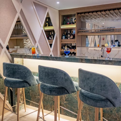 Modern bar interior with three blue velvet bar stools lined up at a marble counter, stocked with various bottles and glasses above and behind the bar.