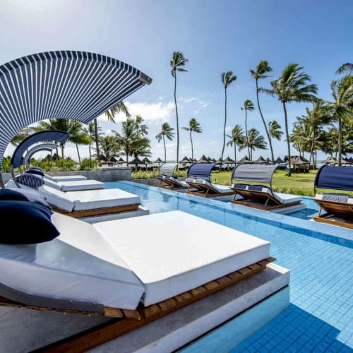 Außenpoolbereich in einem Resort mit modernen weißen Liegen, blauen Kissen und gebogenen Sonnenschutzdächern, umgeben von Palmen und blauem Himmel.