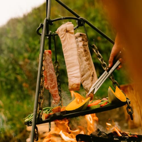 Fleisch und Gemüse, die auf einem hängenden Grillrost über offenem Feuer im Freien gegrillt werden, mit einer Grillzange, die eine Wurst hält.