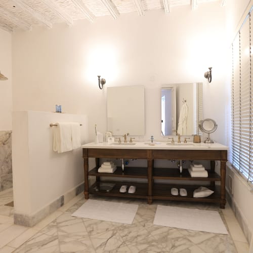 Helles Badezimmer mit Marmorboden, Doppelwaschbecken mit Spiegeln, weißen Handtüchern, begehbarer Dusche und einem Fenster mit Jalousien.