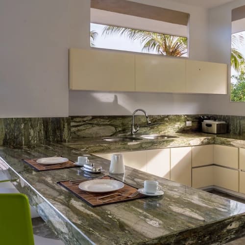 Moderne Küche mit grünen Marmor-Arbeitsplatten und offenem Blick auf Palmen und Wasser durch große Fenster und eine Glastür.