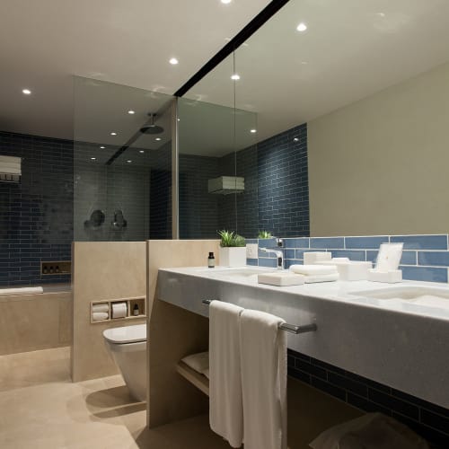 Modern bathroom with blue tiled walls, a glass shower enclosure, a double sink vanity with white towels, and beige flooring.