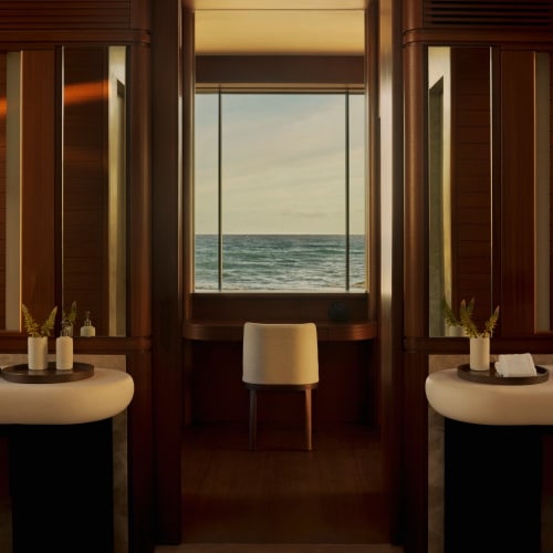 Elegantes Badezimmer mit zwei Waschbecken und einem zentralen Sitzbereich, mit großem Fenster und Blick auf den Ozean bei Sonnenuntergang.