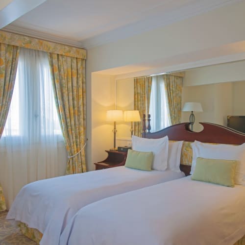 Two neatly made twin beds with white linens and pastel green pillows in a luxurious hotel room, with large windows and floral curtains.