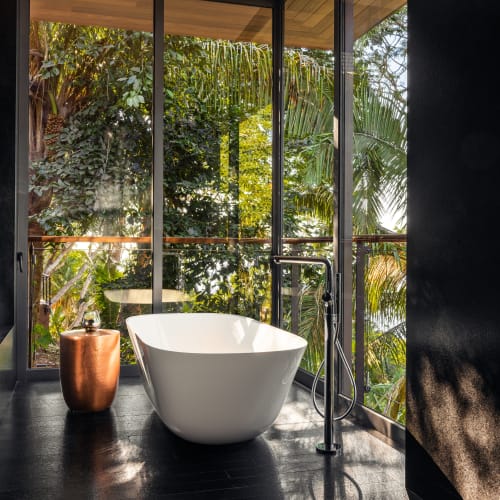 Modernes Badezimmer mit einer freistehenden weißen Badewanne, einem kupferfarbenen Hocker und einem großen Fenster mit Blick auf tropisches Grün.