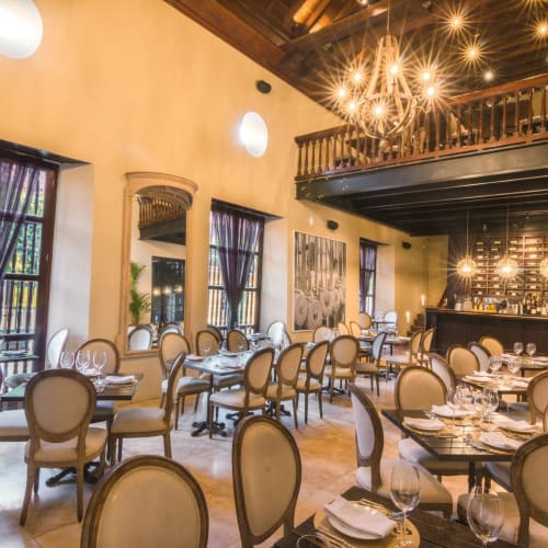 Elegant hotel restaurant with round tables set for dining, large windows with dark curtains, and ornate lighting fixtures.