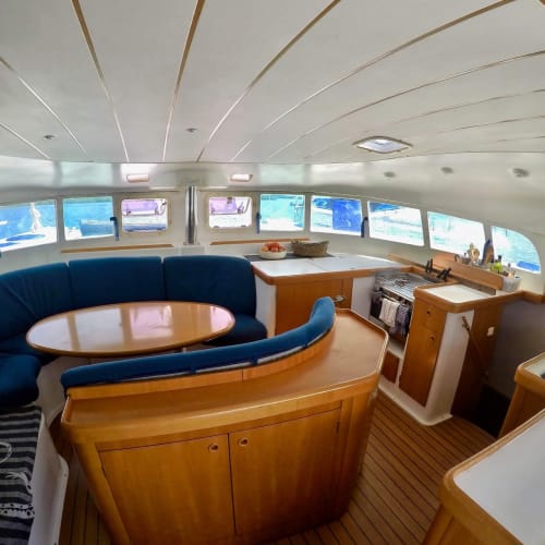 Innenraum einer Yacht-Salon mit einer gebogenen blauen Couch, Holztischen und Arbeitsplatten, und Fenstern, die Wasser zeigen.