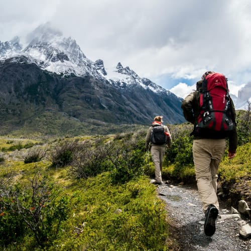 Zwei Wanderer mit großen Rucksäcken wandern auf einem Pfad in einer bergigen Landschaft mit schneebedeckten Gipfeln.