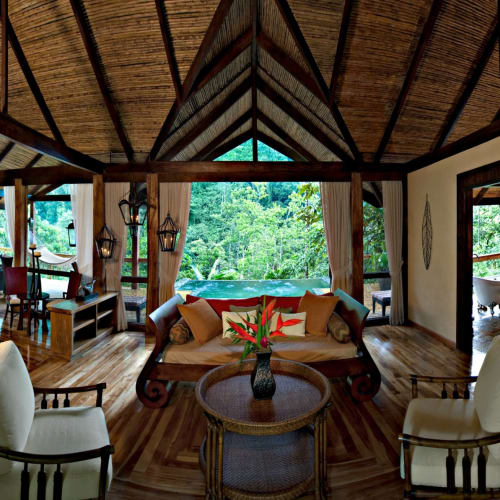 Innenraum eines gemütlichen Wohnbereichs in einer Villa im Pacuare Lodge, mit Holzmöbeln, einem Sofa mit Kissen und großen Fenstern mit Blick auf üppiges Grün.