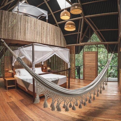 Innenraum eines rustikalen Holzlodges mit einem Himmelbett mit weißen Vorhängen, einer hängenden Hängematte und Holzwänden und -decke mit Pendelleuchten, Blick auf üppiges grünes Laub durch große Fenster und offene Tür.