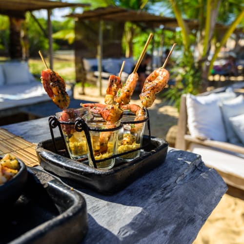 Grilled shrimp skewers served in shot glasses placed on a rustic wooden table at an outdoor beachside setting with palm trees and lounge seating in the background.