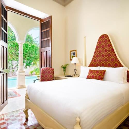 Elegantes Hotelzimmer mit großem weißen Bett und hohem, gemustertem roten und goldenen Kopfteil, zwei Nachttischen mit Lampen und doppelten Holztüren, die auf einen Garten mit Swimmingpool hinausgehen.