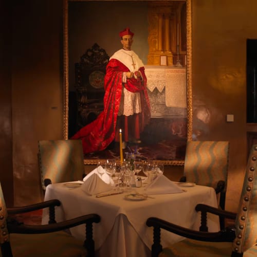 Elegant restaurant dining area with a round table set for a meal, surrounded by upholstered chairs and a large portrait painting on the wall.