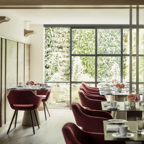 Modernes Restaurantinterieur mit Holzvertäfelung, großen bodentiefen Fenstern mit Blick auf üppiges Grün draußen und Tischen, die mit weißen Tassen, Gläsern und kleinen Blumenarrangements gedeckt sind. Rote Polsterstühle sind ordentlich um die Tische angeordnet und laden zum Verweilen ein.