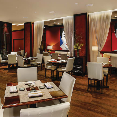 Elegantes Restaurantinterior mit weißen Stühlen, Holztischen, kunstvollen Porträts an den roten Wänden und einer gut bestückten Bar im Hintergrund.