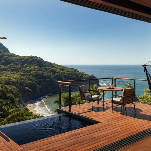 Eine Holzterrasse mit zwei Stühlen, einem kleinen Tisch, einem Infinity-Pool und einem Hängesessel mit Blick auf einen bewaldeten Hügel und das Meer unter klarem Himmel.