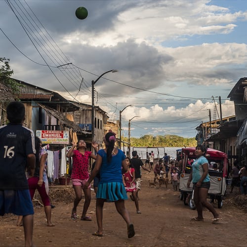 Kinder und Erwachsene spielen Fußball in einer ländlichen Gemeinschaft auf der Straße unter wolkigem Himmel.