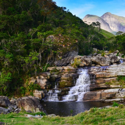 A small waterfall cascades over rocks into a pool surrounded by lush greenery and mountains in the background.