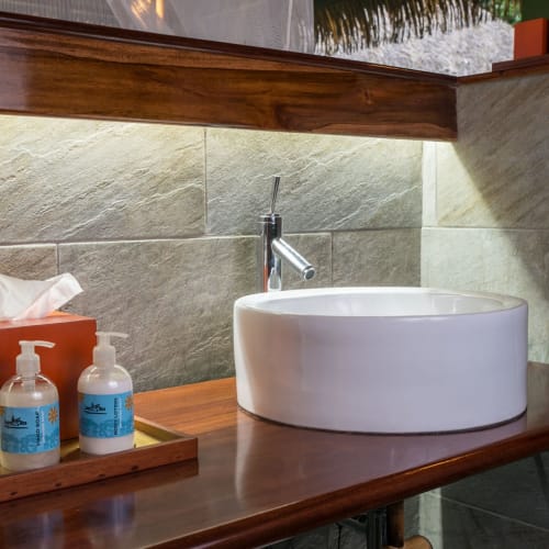 A modern bathroom with a white round basin sink, a wooden countertop, and a faucet, set against textured stone walls with an illuminated mirror above.