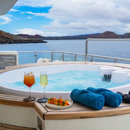 Rundlicher Außenwhirlpool auf dem Deck einer Yacht mit zwei Getränken, einem Teller mit Obst und aufgerollten blauen Handtüchern, mit Blick auf ruhiges Meer und entfernte felsige Inseln unter teils bewölktem Himmel.