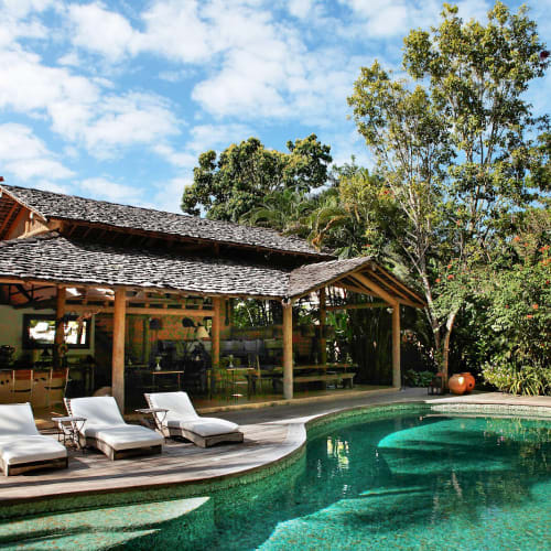 Outdoor swimming pool with three white lounge chairs on a wooden deck next to a large shaded pavilion surrounded by lush green trees under a partly cloudy sky.