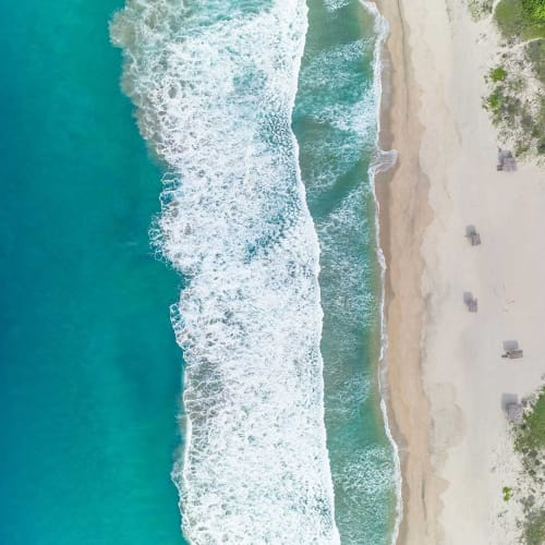 Luftaufnahme einer Küstenlinie mit türkisfarbenem Ozean, der Wellen an einen Sandstrand schlägt, an dem verstreut Strandhütten und grüne Vegetation auf der rechten Seite zu sehen sind.