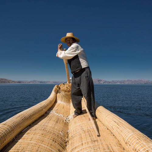 Mann steht auf einem traditionellen Schilfboot und steuert auf einem ruhigen Gewässer unter klarem blauem Himmel, im Hintergrund sind ferne Berge sichtbar.