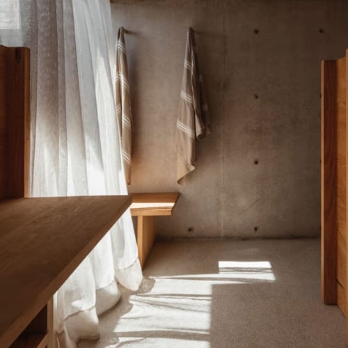 Sonnenlicht, das durch transparente weiße Vorhänge auf ein minimalistisches Badezimmer mit Betonwänden, hölzernen Bänken und zwei aufgehängten Handtüchern fällt.