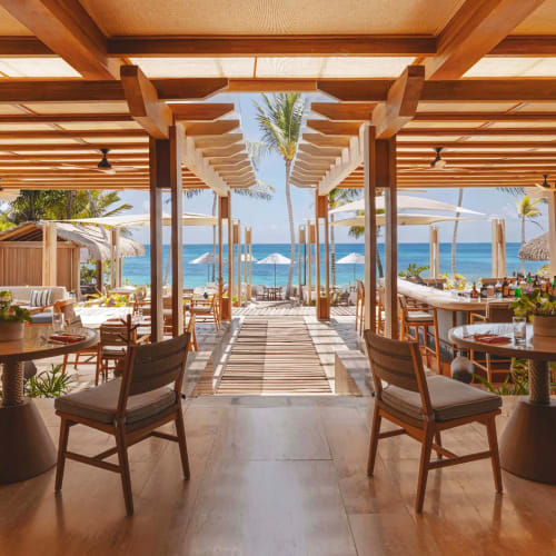 Offener, luxuriöser Bar- und Restaurantbereich mit Holzmöbeln und Blick auf den Strand und das Meer unter einem Holzpergola-Dach.